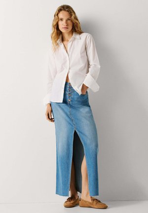 Kvinne iført hvit skjorte med knapper, høy midjet jeansskjørt med frontsplitt, og brune mokkasin-sko, stående mot en ensfarget vegg.