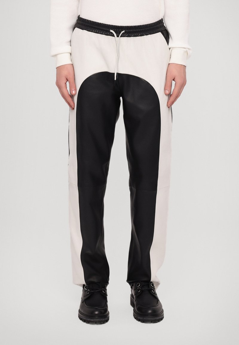 Homme portant un pantalon bicolore noir et blanc avec taille élastique et chaussures noires, debout devant un fond uni.