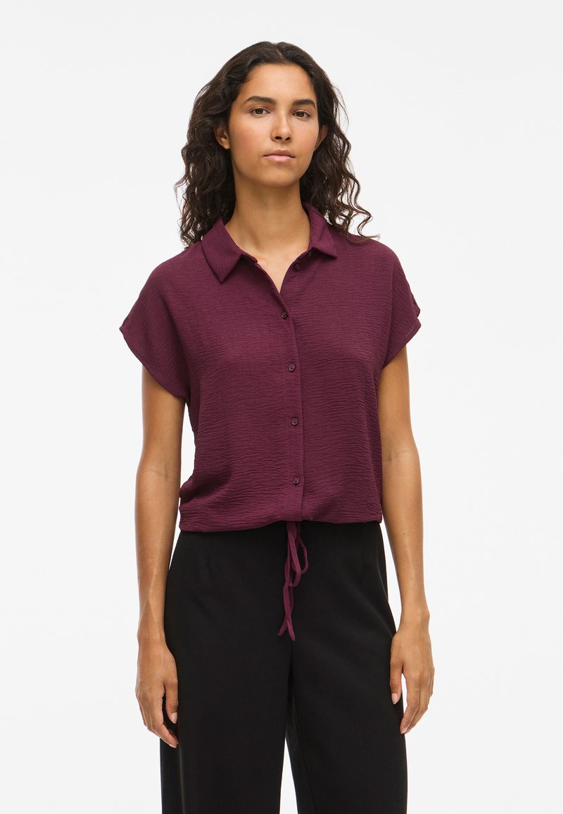 Burgundy short-sleeve shirt with a collar, textured fabric, button closure, and drawstring at the waist, paired with black trousers.