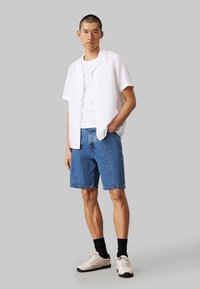 Camisa blanca de manga corta, camiseta blanca debajo, pantalones cortos de mezclilla azul y zapatillas beige con acentos negros. Textura suave, diseño casual.