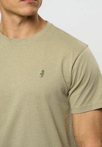 Nærbilde av en person som har på seg en beige t-skjorte med rund hals og et lite mørkegrønt brodert rev logo på brystet.