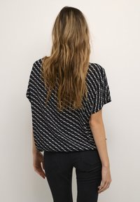 Femme aux cheveux longs et ondulés portant un chemisier noir à motifs et un jean noir, debout face à un fond clair uni.