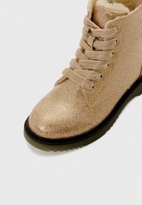 Bottine dorée pailletée avec de grosses lacets beiges, semelle noire et doublure beige duveteuse visible sur le dessus.