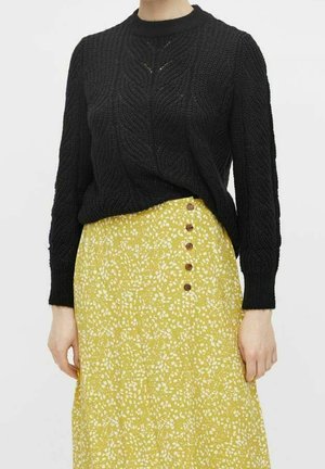 Pull noir en tricot avec un motif texturé, associé à une jupe jaune ornée de motifs floraux blancs et de boutons marron sur le devant.