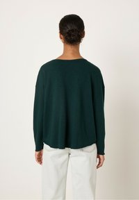 Tumši zaļš pārmērīgs džemperis ar noapaļotu kakla izgriezumu un brīvu piegājienu. Mīksta tekstūra, garas piedurknes un izliekts apakšmala. Saskaņots ar gaišiem biksēm.