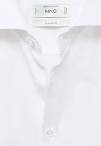 Camisa blanca de botones con corte clásico, que presenta una textura suave, cuello puntiagudo y botones de plástico transparente. La etiqueta dice "MNG THE SUIT COLLECTION".