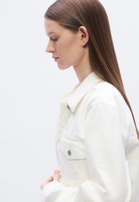 Λευκό τζιν μπουφάν με κλασικό σχέδιο, που διαθέτει μανικοκουμπωτές τσέπες στο στήθος και ομαλή υφή. Γιακάς με δομημένο σχήμα, που ενισχύει τη μορφή του.