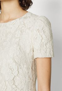 Krajková šaty v off-white barvě s květinovým vzorem, krátkými rukávy a kulatým výstřihem s texturovanými detaily.