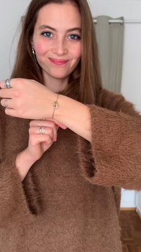 Zlatý náramok so špirálovým vzorom a malými čírymi kameňmi, nosený na zápästí spolu s viacerými striebornými prsteňmi a hnedým chlpatým svetríkom.