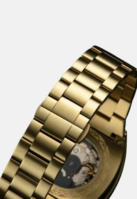 Gouden metalen polshorloge met een geborstelde afwerking, voorzien van een schakelen band en een ronde kast die mechanische beweging toont door een transparante achterkant.