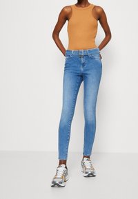 Mujer con pantalones ajustados de talle alto en azul claro con cinturón, top sin mangas color camel y zapatillas blancas-grises, de pie contra un fondo blanco.