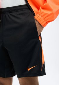 Άνδρας που φοράει μαύρο αθλητικό σορτς Nike με πορτοκαλί πλαϊνές λωρίδες και λογότυπο, συνδυασμένο με πορτοκαλί μπουφάν, με το χέρι στην τσέπη.