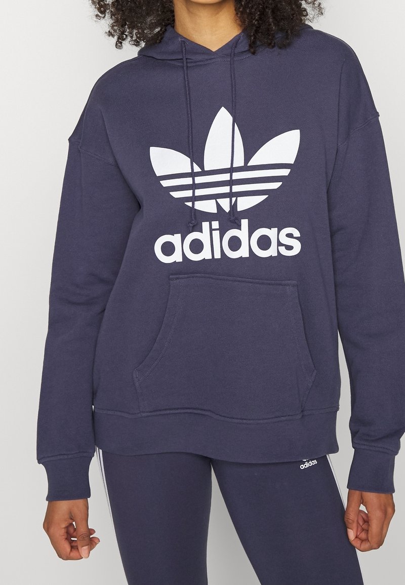 Granatowa bluza z kapturem z białym logo Adidas Trefoil, sznureczkami, kieszenią kangurką i ściągaczami oraz wykonana z miękkiego, fakturowanego materiału.