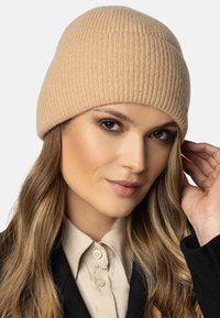 Beige ribbelgebreide beanie muts met een zachte textuur. De muts heeft een aangesloten pasvorm en een ronde bovenkant, en wordt iets scheef op het hoofd gedragen.