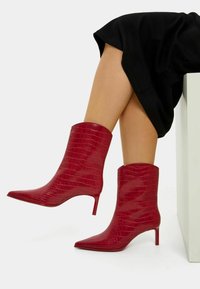 Red ankle boots made of glossy, croc-embossed leather with a pointed toe and a slim heel. The design is sleek and fitted around the ankle.