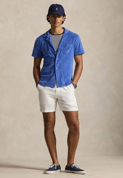 Νεαρός άνδρας με μπλε καπέλο, μπλε πουκάμισο με κοντά μανίκια και κουμπιά, ριγέ μπλουζάκι, λευκά σορτς, μπλε αθλητικά παπούτσια, στέκεται με τα χέρια στις τσέπες σε απλό φόντο.