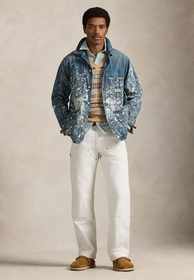 Άνδρας στέκεται με τα χέρια στις τσέπες, φοράει ένα τζιν μπουφάν με πιτσιλιές μπογιάς, πουλόβερ χωρίς μανίκια με μοτίβο, ριγέ πουκάμισο, λευκό παντελόνι και καφέ παπούτσια.