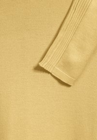 Senapsgul stickad tröja med en jämn textur, ribbad manschett och en kroppsnära design. Materialet är mjukt och elastiskt.