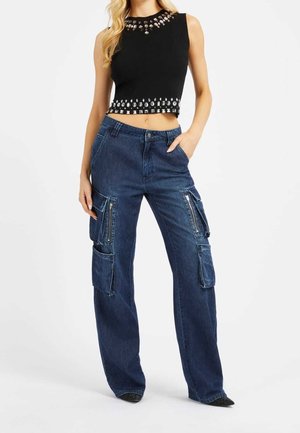 Vrouw draagt een zwart mouwloos crop top met zilveren studs en losse donkerblauwe cargobroek met meerdere zakken en ritsen.