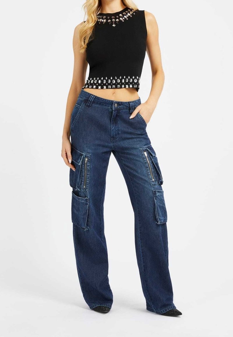 Femme portant un crop top noir sans manches avec des clous argentés et un jean cargo bleu foncé ample avec plusieurs poches et fermetures éclair.