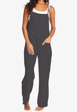 Nainen pukeutuneena löysiin mustiin, napillisiin haalareihin, joista toisessa tasku ja valkoinen toppi alla.