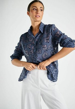 Doorzichtige marineblauwe blouse met donker bloemmotief, knoopsluiting aan de voorkant en opgerolde mouwen, gecombineerd met witte broek met wijde pijpen.