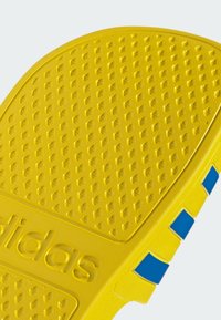 La suela de goma amarilla presenta una superficie texturizada con pequeños patrones elevados, acentos azules y el logo de "adidas" estampado de manera prominente.