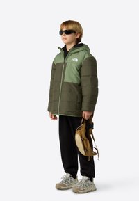 Zöld és sötétzöld kabát kapucnival, elöl cipzáras és logóval. Fekete nadrág és bézs hátizsák. Világosszürke cipők díszítésekkel.