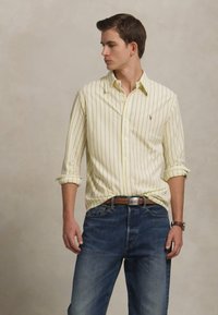 Κίτρινο πουκαμίσα με ρίγες και διπλωμένα μανίκια, σε συνδυασμό με μπλε τζιν παντελόνι. Διαθέτει ένα μικρό λογότυπο στην τσέπη του πουκαμίσου.