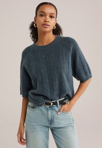 Suéter corto de manga corta de textura azul oscuro con un escote redondeado, combinado con unos vaqueros de lavado claro y un cinturón negro con hebilla dorada.