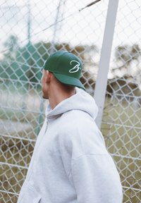Cappello da baseball verde con un logo bianco in corsivo "B", indossato al contrario sopra un felpa grigia, in piedi vicino a una recinzione in catena.