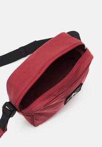 Borsa a tracolla rossa in tessuto con apertura con cerniera, tracolla nera regolabile e una piccola etichetta del logo sul lato. Texture liscia e design minimalista.