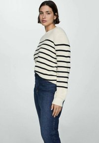 Maglione crema con righe orizzontali nere, scollo rotondo e polsini a costine, abbinato a jeans blu scuro. Tessuto morbido.