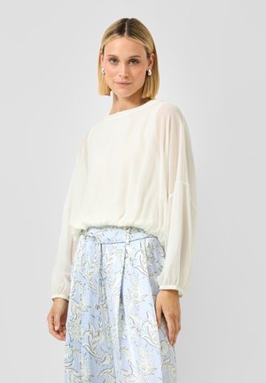 Frau mit kurzen blonden Haaren, die Perlenohrringe, eine weiße Langarmbluse und einen hellblauen, geblümten Rock mit gebundenem Taillengürtel trägt.