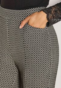 Hand in der Tasche von schwarz-weißen gemusterten Hosen, kombiniert mit einem schwarzen Spitzenärmel und einem gerippten Oberteil.