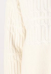 Pull crème en maille torsadée avec des détails côtelés, présentant un motif texturé et un tissu doux. Vue rapprochée de la section de la manches.