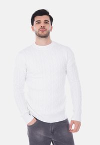 Maglione bianco a maglia con maniche lunghe, collo rotondo e polsini a coste, abbinato a jeans grigio scuro, creando un effetto sovrapposto.