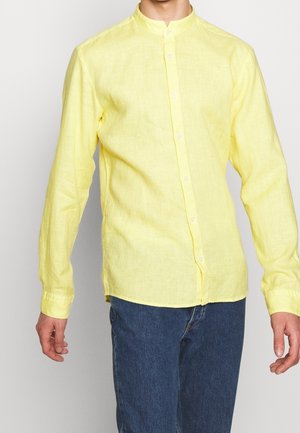 Mężczyzna ubrany w żółtą lnianą koszulę z długim rękawem i stójką oraz niebieskie dżinsy, stojący z rozluźnionymi ramionami.