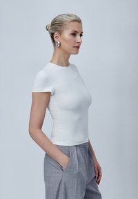 Camiseta ajustada de manga corta blanca, hecha de tela suave, con cuello redondo, combinada con pantalones ajustados de color gris claro, con la mano en el bolsillo.
