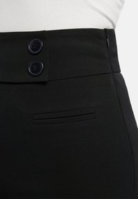 Primer plano de pantalones negros de cintura alta con dos grandes botones, cremallera lateral y un pequeño bolsillo horizontal con abertura por encima de la cadera.