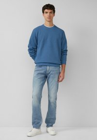 Niebieski teksturowany sweter z okrągłym dekoltem, zestawiony z jasnoniebieskimi dżinsami i białymi trampkami. Neutralne tło.