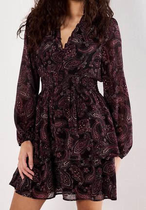 Vrouw die een knielange zwarte jurk draagt met roze paisleypatronen, lange mouwen en een getailleerde taille, staand tegen een effen achtergrond.