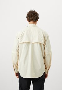 Camicia beige chiaro con dettaglio di yoke sul retro, maniche lunghe e polsini abbottonati; realizzata in un tessuto morbido con una finitura leggermente strutturata.