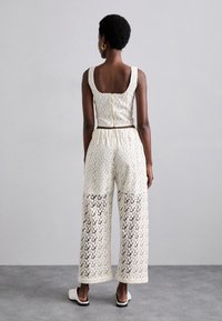 Hvit blonde crop topp med firkantet rygg og gullglidelås, parret med vide blondebukser med mønstrede utskjæringer, båret med slip-on sko.