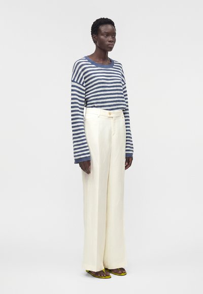 Γυναίκα φοράει μπλε και λευκό ριγέ πουλόβερ με μακριά μανίκια, λευκό παντελόνι με ψηλή μέση και φαρδιά μπατζάκια, και πράσινα ανοιχτά σανδάλια, στέκεται μπροστά σε γκρι φόντο.