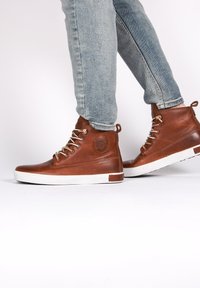Scarpe alte in pelle marrone con suola in gomma bianca, occhielli in metallo e un logo circolare sul lato. Indossate con jeans blu.