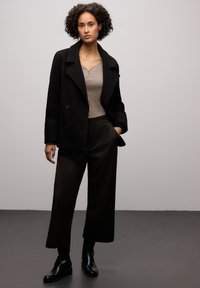 Cappotto nero doppiopetto sopra una maglia beige, abbinato a pantaloni larghi scuri e strutturati. Stivaletti neri completano il look.