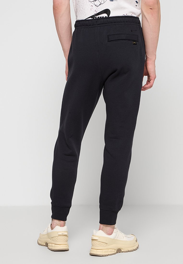 Mann in schwarzen, schmal geschnittenen Jogginghosen und off-white Sneakers, vor einem schlichten weißen Hintergrund stehend.