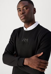Felpa nera con scollo rotondo, caratterizzata da un logo testurizzato al centro. Il modello indossa una camicia bianca con colletto sotto.