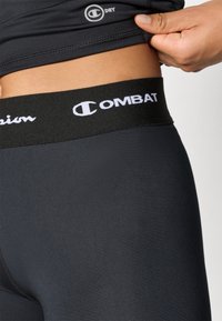 Svarta sporttights med en slät yta och en svart midja som har vita tryckta logotyper för "Champion" och "COMBAT".